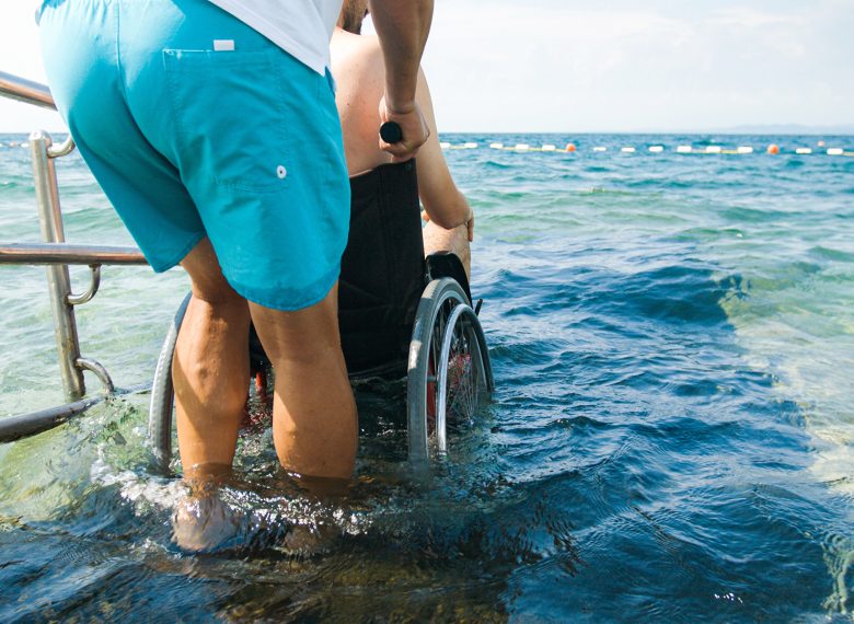 Disabled man at beach swimming on a wheelchair with assistance help on an accessible ramp.
